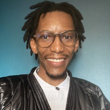 African male with short dreadlocked hair, wearing reading glasses and smiling.